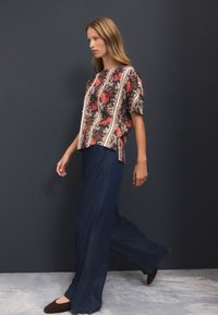 Blouse à manches courtes à motif floral en noir, rouge et crème, associée à un pantalon ample bleu foncé, portée avec des chaussures plates marron foncé.