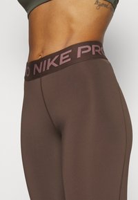 Hnědé legíny Nike Pro s širokým pasem, na kterém je nápis "NIKE PRO" v světle růžové barvě. Hladká, elastická látka s vyztuženými švy.