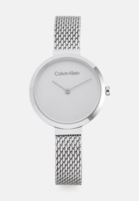 Montre bracelet en maille argentée avec un cadran rond, présentant un cadran blanc minimaliste et un logo subtil. Accents en métal lisse et poli.