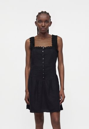 Γυναίκα με πλεγμένα μαλλιά που φοράει μαύρο αμάνικο φόρεμα με κουμπιά μπροστά και δαντέλα στα τιράντες και το ντεκολτέ, μπροστά σε απλό φόντο.