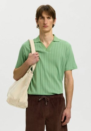Jeune homme portant un polo vert clair à manches courtes et un pantalon marron, tenant un sac beige sur l'épaule gauche, debout devant un fond uni.