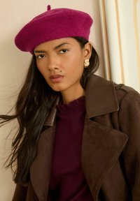 Sieviete ar gariem tumšiem matiem valkā tumši rozā bereti, zelta auskarus, koraļļkrāsas topu un tumši brūnu mēteli, skatoties tieši priekšā.