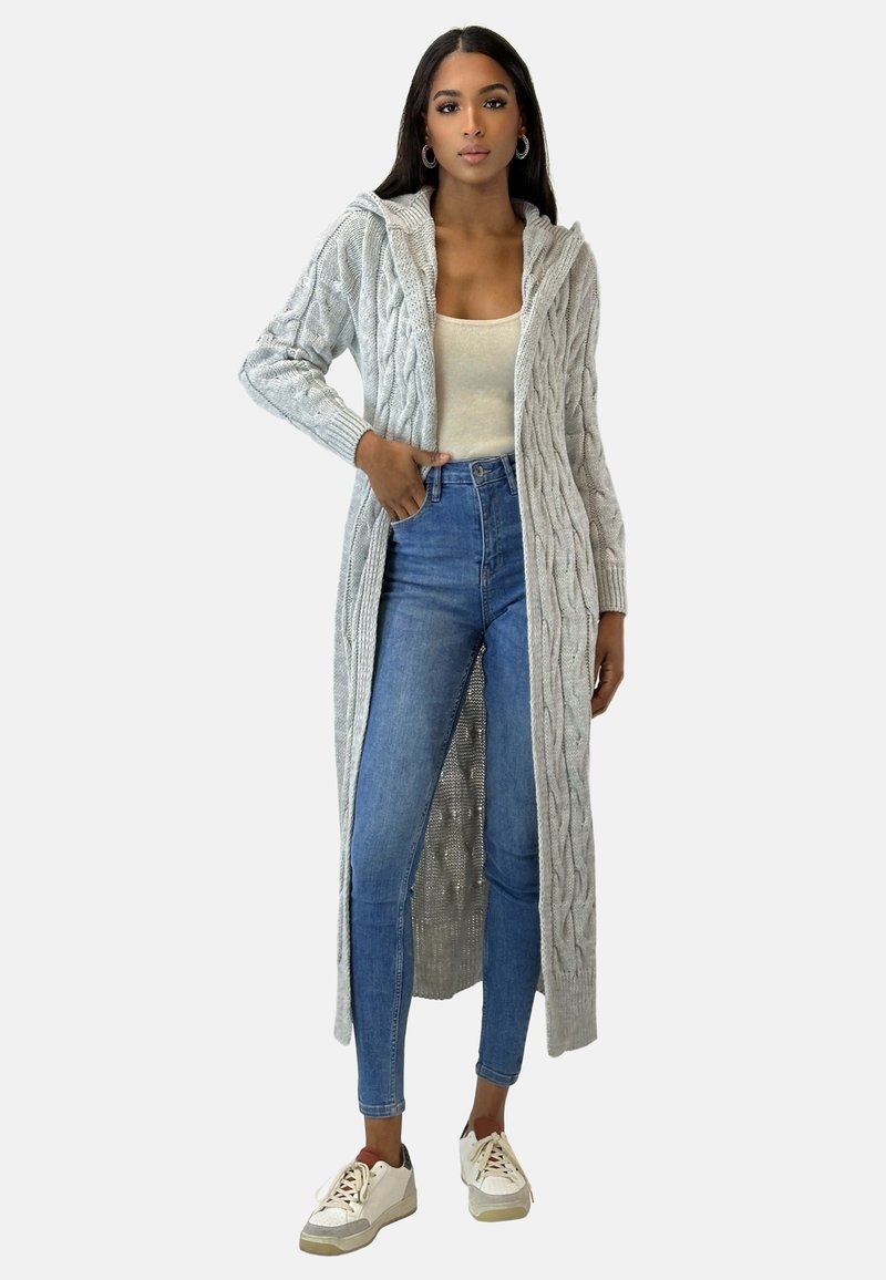Langer grauer Strickcardigan mit Zopfmuster, offener Vorderseite und Kapuze. Getragen über einem hellen Oberteil und hochtaillierten blauen Jeans mit Sneakers.