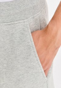 Szare spodnie dresowe z prążkowanym pasem, bocznymi kieszeniami oraz miękką teksturą, skupione na obszarze kieszeni i dłoni spoczywającej w środku.