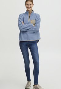 Ljusblå fleece-tröja med halv dragkedja, beige bröstficka och ribbade ärmslut, matchad med mörka tajta jeans och grå sneakers.