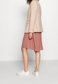 Blazer beige su misura con bottoni dorati, abbinato a una gonna rosa fluida e sneakers bianche, che creano una silhouette rilassata e stratificata.