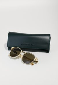 Lacoste prillid, millel on läbipaistev raam ja pruunid läätsed, tumeroheline kunstnahast karp, millel on Lacoste logo.