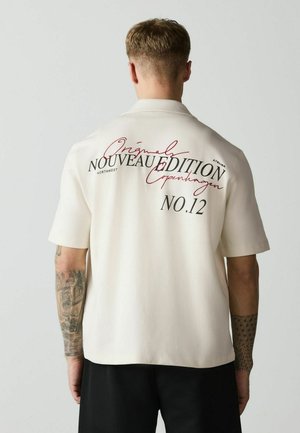 Muž s krátkymi vlasmi a tetovaniami nosí biele tričko s krátkym rukávom s čiernym a červeným textom na chrbte "Original Nouveau Edition Copenhagen No.12".