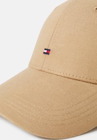 Khaki baseball-hattu, joka on valmistettu teksturoidusta kankaasta. Edessä on pieni brodeerattu logo, jossa on punaisia, valkoisia ja sinisiä raitoja.