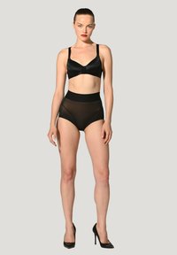Zwarte satijnen bralette met verstelbare bandjes, gecombineerd met hoge sheer zwarte slips. Draagt zwarte stiletto hakken.