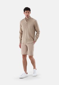Beige Langarmhemd mit Knopfverschluss und Tasche, kombiniert mit beige Shorts und weißen Sneakern, vor einem schlichten Hintergrund stehend.