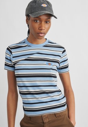 Νέα γυναίκα που φοράει μαύρο καπέλο, μπλε και μαύρη ριγέ μπλούζα και καφέ παντελόνι μπροστά σε ένα απλό φωτεινό φόντο.