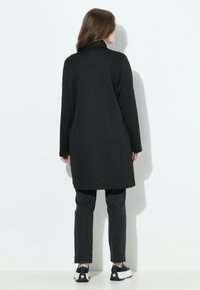 Mujer de espaldas vistiendo un abrigo negro largo, pantalones negros y zapatillas negras con suelas blancas y detalles de estrellas, de pie contra una pared blanca.