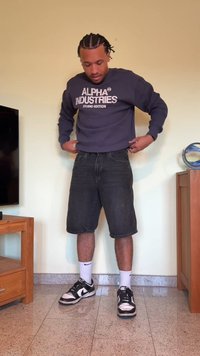 Sweatshirt azul-marinho com o texto "ALPHA INDUSTRIES", calções de ganga pretos e sapatilhas Nike pretas. O modelo está em pé num chão de azulejos ao lado de móveis.