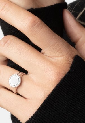 Rosaguld ring med et cirkulært centrum, der har en glat hvid overflade og en tekstureret sølvkant, båret på en hånd med en sort sweater.