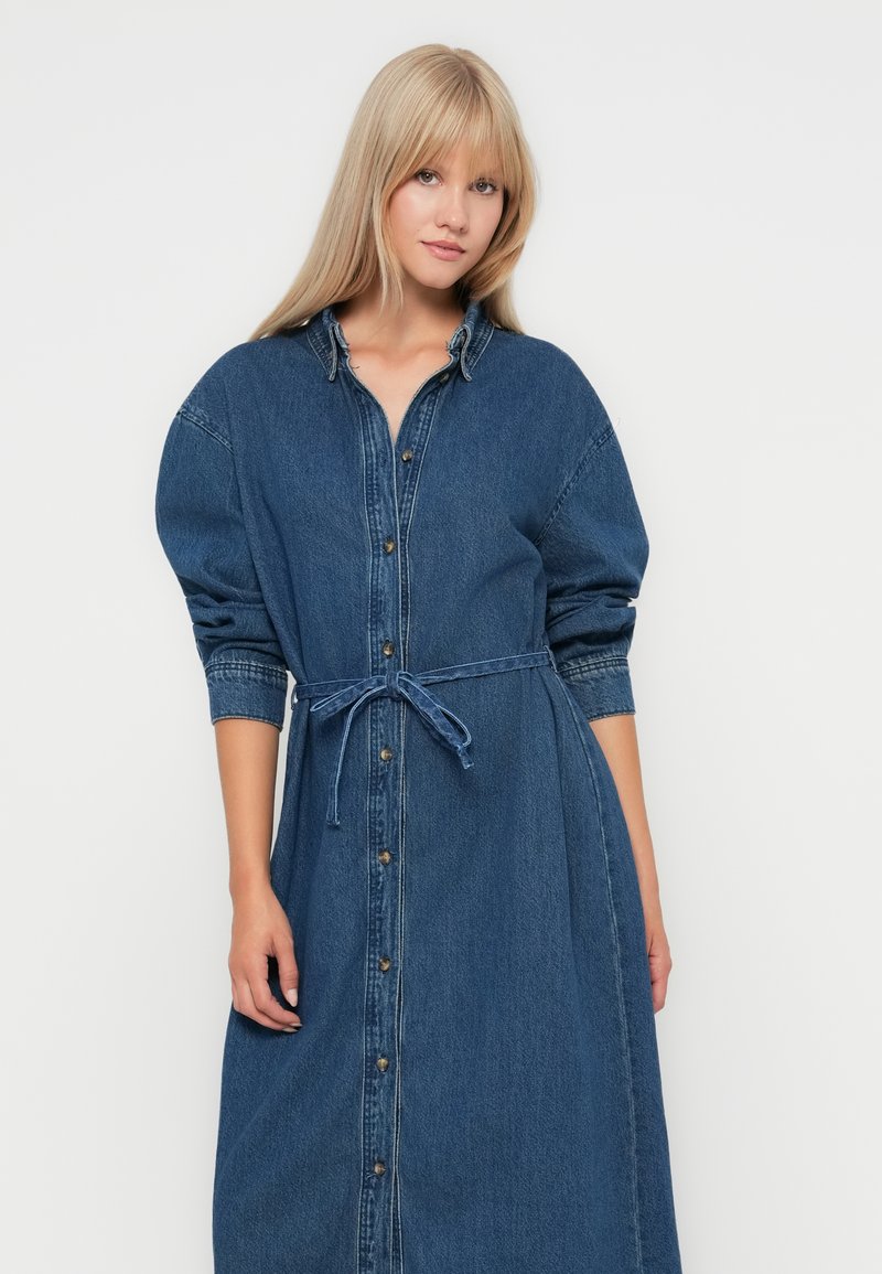 Sukienka koszulowa z denimu, długie rękawy, z przodu na guziki, wcięcie w talii z dopasowanym paskiem, w średnim odcieniu niebieskiego z lekko wyblakłą teksturą.