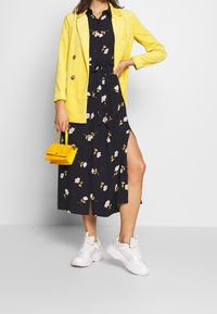Gelber Cord-Blazer, schwarzes Blumenkleid mit Seitenschlitz, weiße Sneakers und kleine gelbe Handtasche. Verschiedene Texturen und lebendige Farben.
