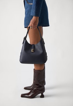 Persona che indossa un vestito blu e stivaletti con tacco marrone scuro, tenendo una borsa nera in pelle con chiusura dorata al lato.