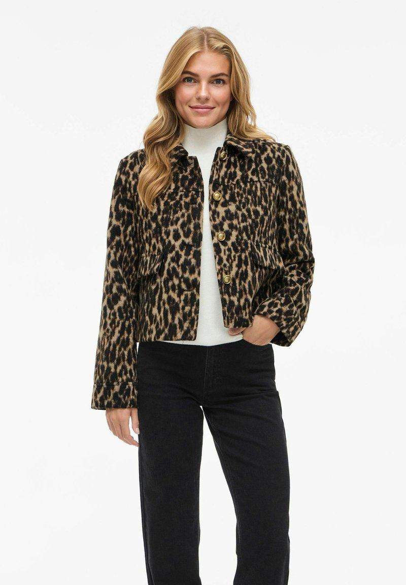 Giacca con stampa leopardata e bottoni dorati, design cropped e due tasche frontali. Indossata sopra una maglietta chiara e abbinata a pantaloni neri.