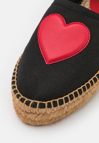Black espadrille with a woven jute sole and a red leather heart appliqué on the upper. Features contrasting stitching around the sole.