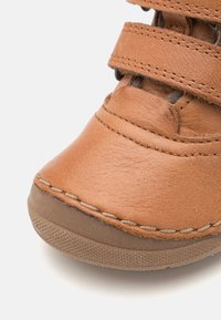 Chaussure en cuir tan avec un bout arrondi, des bandes Velcro, des coutures visibles et une semelle en caoutchouc texturée. Elle présente une surface lisse et un léger éclat.