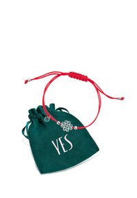 Czerwony pleciony sznurek bransoletki z srebrnym charms o kształcie kwiatu i dwoma srebrnymi koralikami, zaprezentowany z zielonym welurowym woreczkiem oznaczonym napisem "TAK".