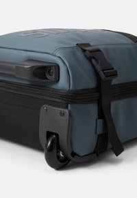 Valise à roulettes avec un extérieur bleu doux, une base noire et des accents texturés ; elle dispose d'une fermeture éclair, d'une poignée et de roulettes qui roulent en douceur.