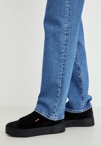 Black suede sneakers with a thick black sole, featuring red logo accents, paired with blue denim jeans, creating a casual look.