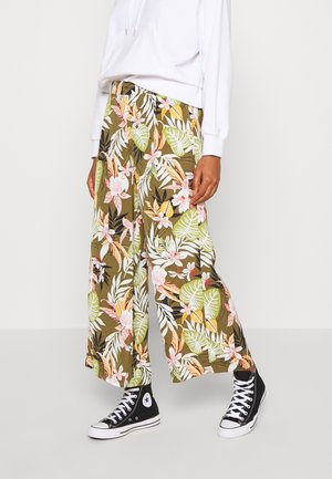 Personne portant un pantalon à jambes larges à imprimé floral avec des feuilles et des fleurs vertes, roses et blanches, un sweat à capuche blanc et des baskets montantes noires.
