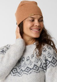 Beanie lavorato a maglia marrone con etichetta, abbinato a un maglione grigio chiaro con un motivo geometrico in grigio scuro. Texture liscia, vestibilità aderente.