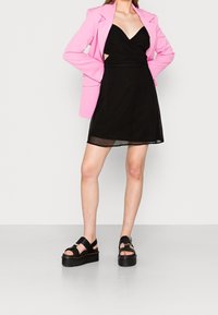 Svart omslagsklänning med djup utringning, kombinerad med en ljusrosa blazer. Outfitten har också tjocka svarta platåsandaler.