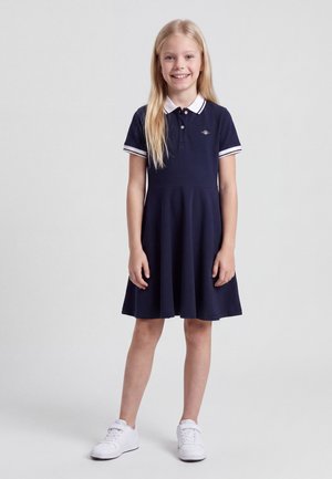 Fată tânără blondă zâmbitoare, stând în picioare, purtând o rochie polo albastru închis cu guler și manșete albe și adidași albi pe un fundal deschis.