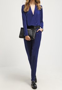 Mono azul marino con diseño envolvente, mangas largas y cintura ajustada, combinado con un bolso negro de mano y zapatos negros brillantes.