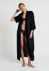 Robe en satin noir avec des manches kimono, devant ouvert et accents en dentelle. Portée sur un soutien-gorge en dentelle noire et un bikini assorti. Texture lisse et brillante.