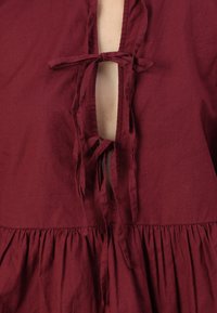 Burgundi spalvos medvilnės viršus su iškirpte, pasižymintis surišamais akcentais ir surinkta liemeniu, užtikrinantis laisvą prigludimą.