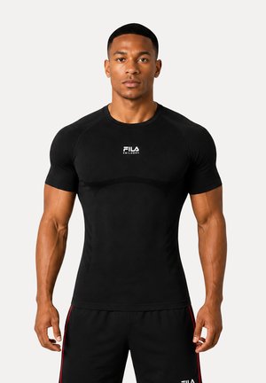 Muskelöser Mann, der nach vorne schaut, trägt ein eng anliegendes schwarzes FILA kurzärmeliges Sportshirt und passende schwarze Shorts mit roten Streifen.