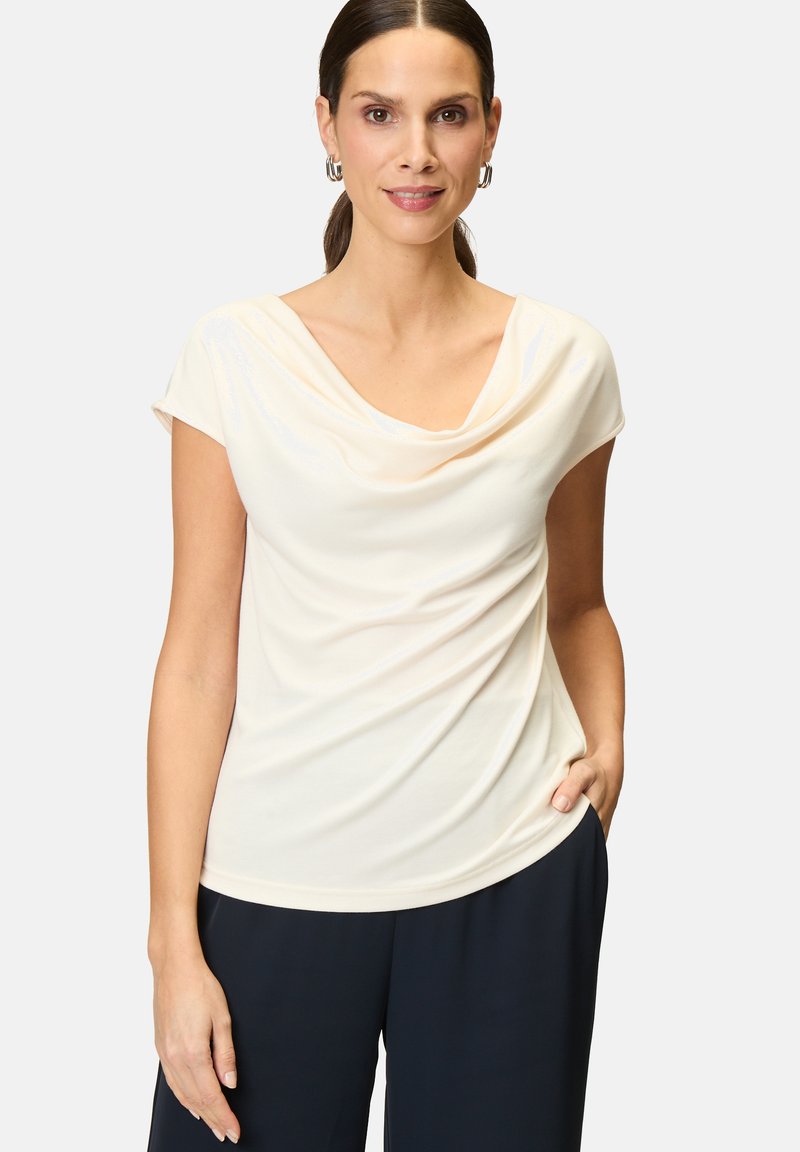 Cremfarbener Top mit Cowl-Neck aus weichem Material, mit kurzen Ärmeln und drapiertem Design. Kombiniert mit marineblauen Hosen; ein subtiler Glanz verleiht Textur.