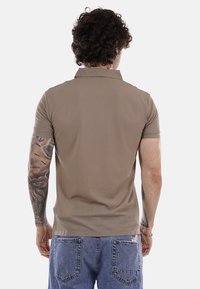 Hombre con cabello rizado que lleva una camisa polo de manga corta beige con textura y jeans de mezclilla azul, mostrado de espaldas.