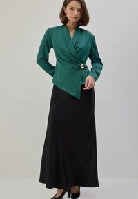 Mujer con blusa cruzada verde esmeralda con cierre dorado y falda larga negra, posando frente a un fondo liso gris claro.