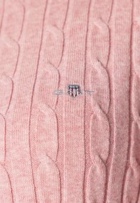 Tissu tricoté torsadé rose avec un petit logo brodé représentant un écusson avec des étoiles et des bandes au centre.