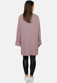 Vrouw staand met de rug naar de camera, gekleed in een lange mauve gebreide cardigan, zwarte legging met leren look en witte sneakers, tegen een effen achtergrond.