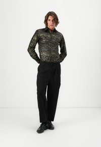 Schwarz und gold gemusterte Button-Up-Hemd mit Wellenmuster, kombiniert mit lockeren schwarzen Hosen und schwarzen Slippern. Glatte Textur.