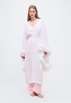 Žena oblečená v priesvitnom bielom župane s nadýchanými manžetami cez ružové pyžamo, stojaca bosá na bielom pozadí.