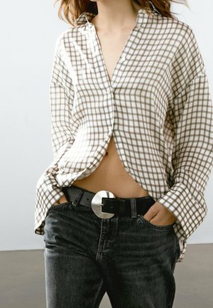 Femme portant une chemise ample beige et blanche à carreaux légèrement rentrée dans un jean foncé avec une ceinture noire et une boucle circulaire argentée.