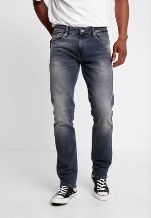 Uomo che indossa jeans slim fit grigio scuro sbiaditi, sneakers bianche e nere e una maglietta bianca, in piedi davanti a uno sfondo semplice.