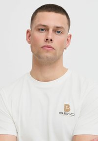 Balta kokvilnas T-kreklu ar apaļu kakla izgriezumu, ar mazu brūnu logo kreisajā krūtīs, kas lasa "BLEND COPENHAGEN". Īsas piedurknes.