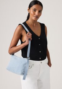 Γυναίκα που φοράει μαύρο αμάνικο τοπ με κουμπιά και λευκό παντελόνι, κρατώντας μια γαλάζια τσάντα ώμου με φαρδιά τιράντα πάνω από τον δεξί της ώμο.