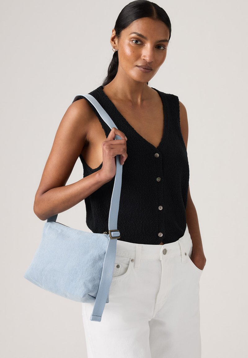 Γυναίκα που φοράει μαύρο αμάνικο τοπ με κουμπιά και λευκό παντελόνι, κρατώντας μια γαλάζια τσάντα ώμου με φαρδιά τιράντα πάνω από τον δεξί της ώμο.