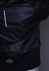 Veste de course légère noire avec une finition brillante, présentant une bande verticale blanche et un t-shirt noir en dessous avec un logo textuel.