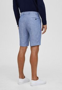 Pantaloni corti di lino azzurro chiaro con una texture liscia, dotati di una tasca posteriore con bottone. Indossati con sneakers bianche, mostrano un aspetto casual.